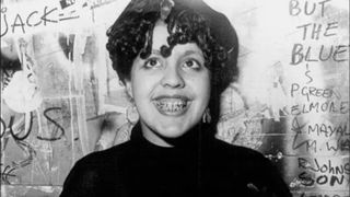 A black and white photo shows punk icon Poly Styrene standing against a wall covered in grafitti