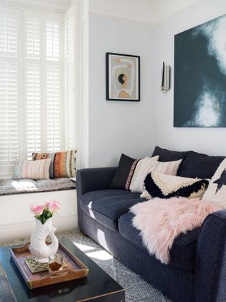 a light bright living room with blue sofa and pink and white cushions, and dark coffee table with a tray and flowers on top, and a window seat with cushions, and white shutters on the windows