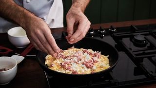 Hands adding ham hock to omelette in skillet
