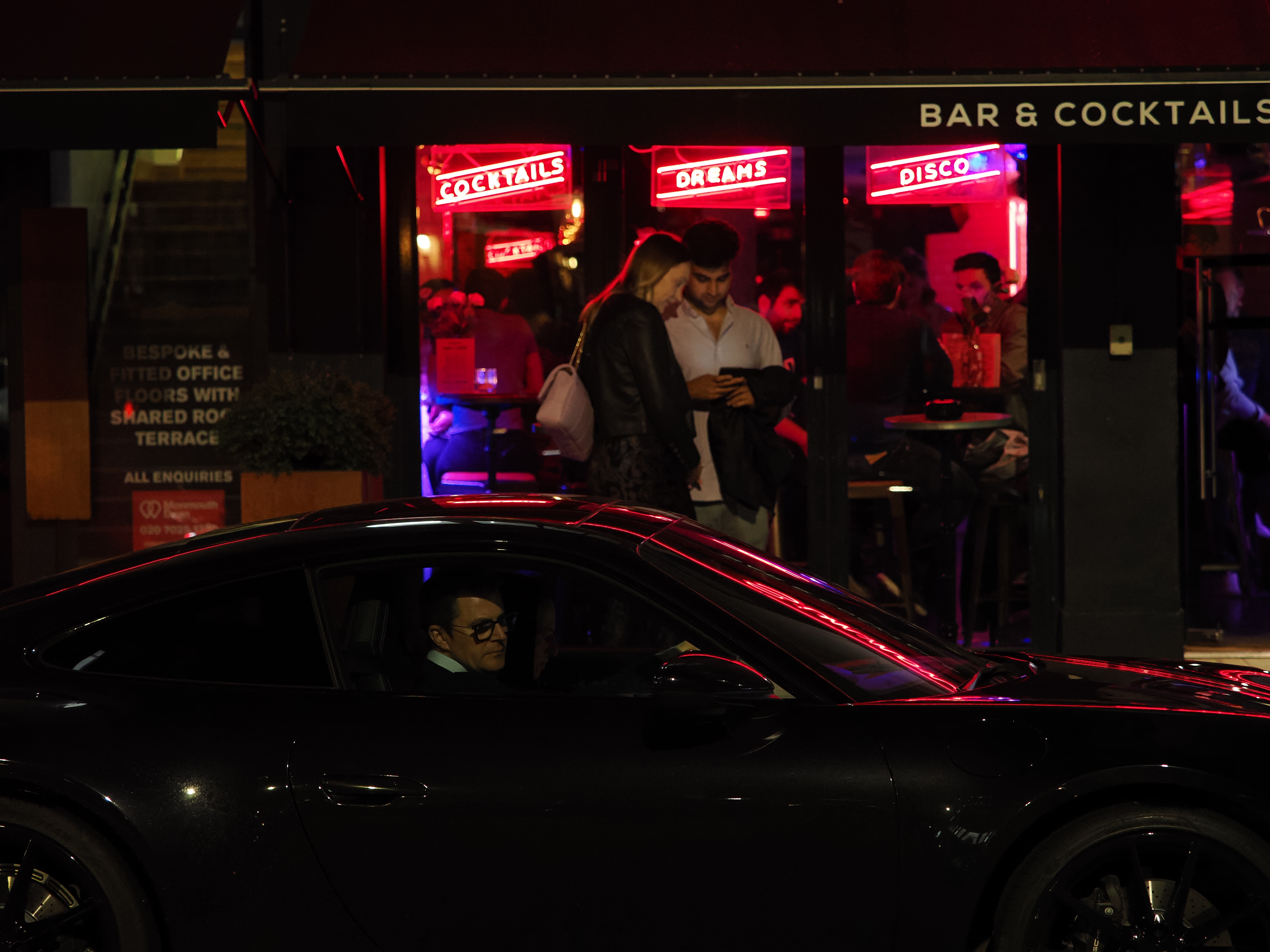 Two people standing outside a bar at night behind a taxi