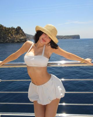 Kylie Jenner posing on a yacht in front of the ocean wearing a white bikini top, white bubble mini skirt, and straw hat.