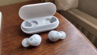 A white pair of Sony WF-C700N in-ear headphones on a wooden tabletop. The open case is positioned behind them.