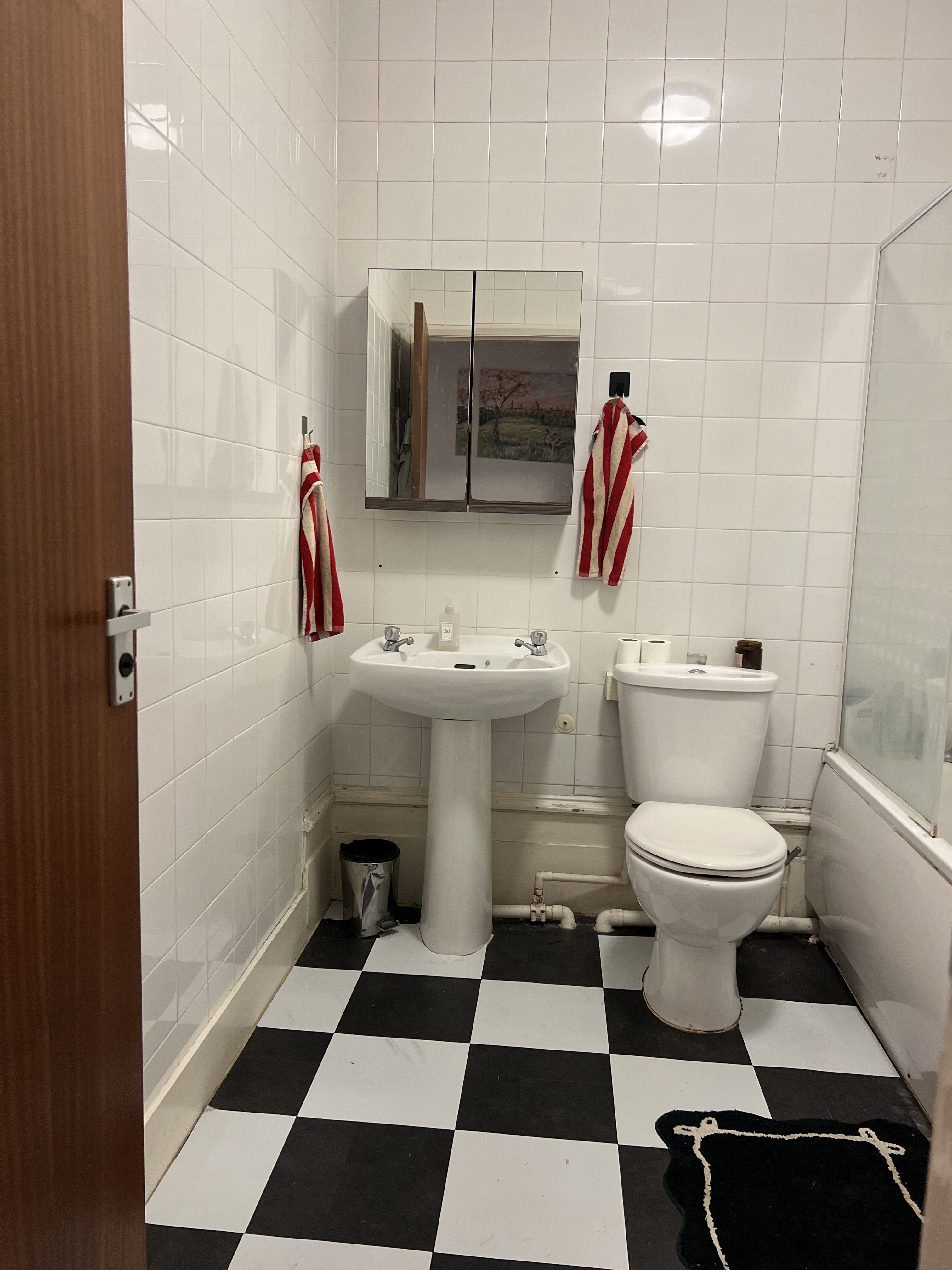 A bathroom with black-and-white checkerboard floors with all-white tiled walls, and all white appliances. There are two red and beige striped towels hanging by the sink.