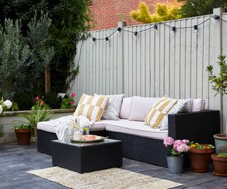 Grey painted fence hung with festoon lights and an outdoor corner sofa in front with cushions