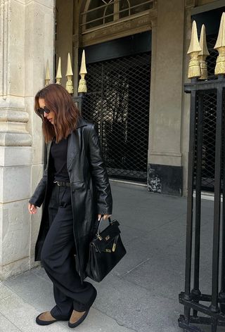 how to wear Chelsea boots is shown in a photo of a woman in pairs wearing a long black leather coat over a black sweater with black trousers, a black handbag, and brown suede chelsea boots