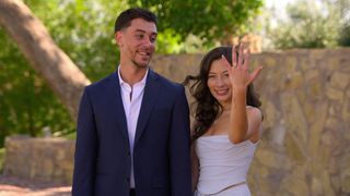 Caleb stands next to Mariah as she holds up her engagement ring on her left hand, while standing in front of a stone wall outdoors, in 'The Ultimatum.'