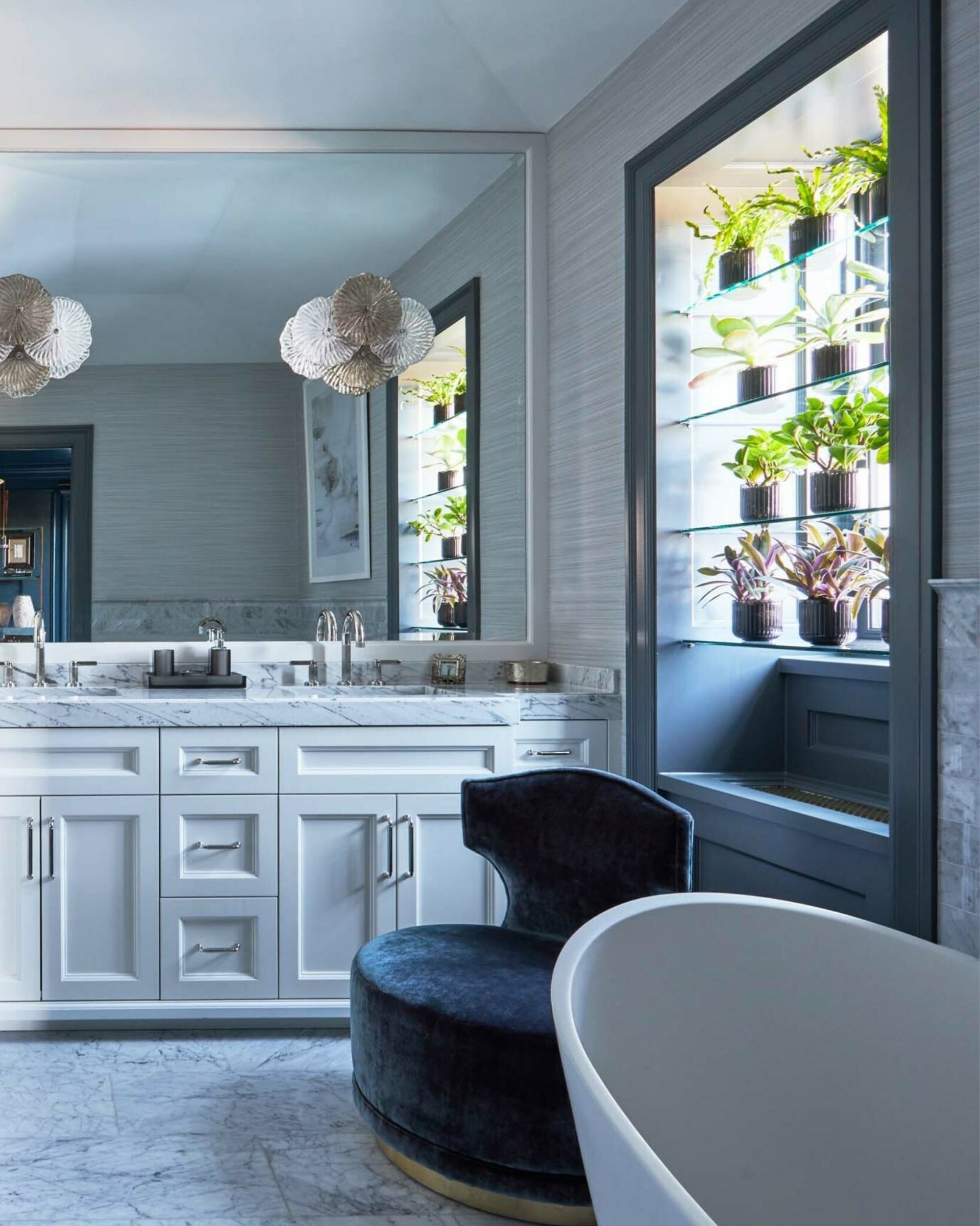 A large, luxurious, and gray marble bathroom with a large window that has glass shelves with plant pots lined on them.