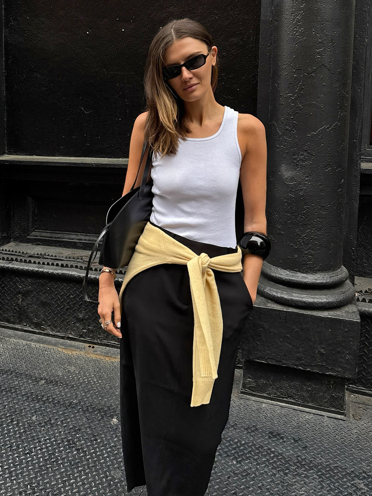 NYC style influencer Coco Schiffer poses on a sidewalk wearing black rectangular sunglasses, a white tank top, black minimal shoulder bag, a large black cuff bangle bracelet, yellow sweater tied around the waist, and a black midi skirt.