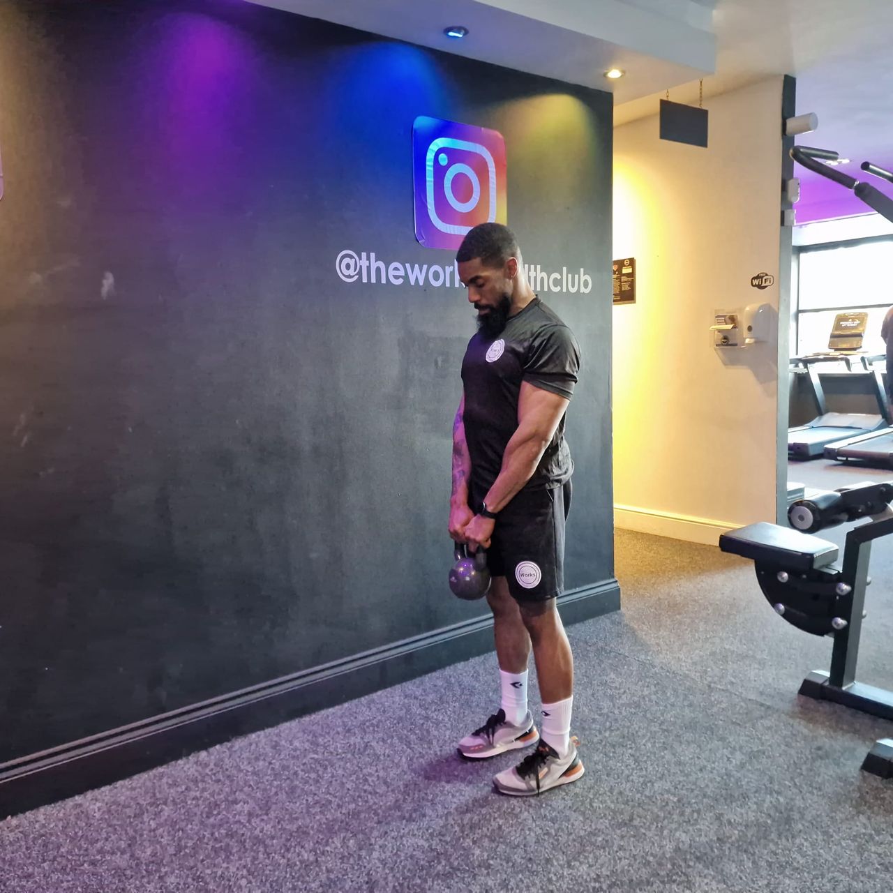 Renel Scarlett performs a kettlebell deadlift at The Works Health Club gym. He is standing, with his arms hanging straight down and holding a kettlebell at his thighs. Behind him we see empty treadmills.