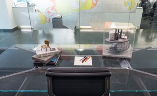 Black chair and glass desk with books, a pad, pens and a miniature architectural model in a clear case. The office partition wall made of glass features a world map