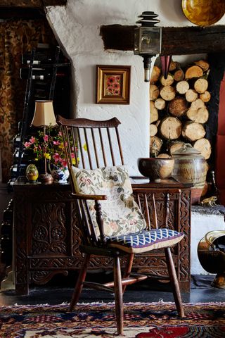 wooden chair by the fireplace, antique sideboard, vintage rug, log pile, small vase of flowers, artwork