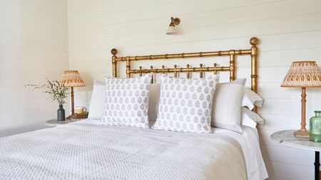 Narrow bedroom ideas can still be so stylish. A narrow bedroom with a gold metal bed frame with gray textured bedding and two white and blue circle aptterned throw pillows, with a nightstand to the left with a curved blue vase and brown table lamp