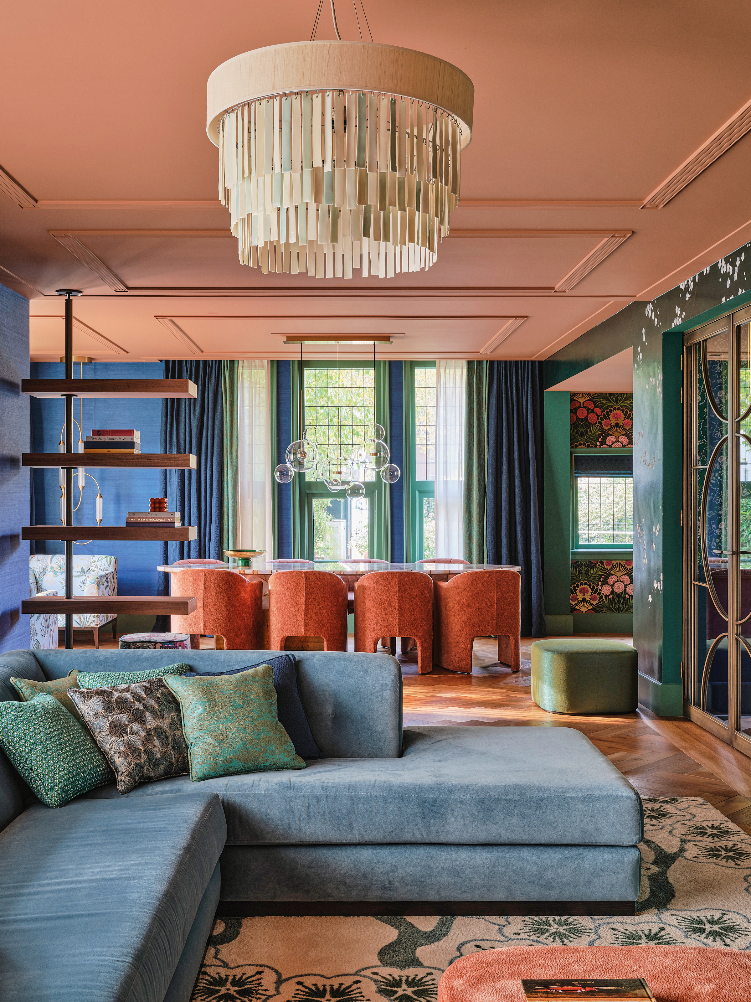 Living and dining area with green and blue walls, salmon pink ceiling, blue corner sofa, coral dining chairs and a cream chandelier