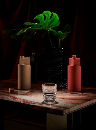 Three ceramic bottles of OAX Original mezcal in different colours on a marble table next a crystal glass and black vase with a green plant