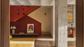 A living room with white walls, concrete pillars, a mustard bench, and a red enclosed staircase that zig-zags in the background