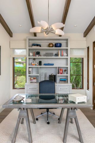 Home office with neutral decor, glass and wood desk, desk chair, shelving and pendant light