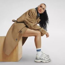 Sydney McLaughlin-Levrone sat on wooden block wearing trench coat, blue-striped socks and New Balance 9060 in grey and white.