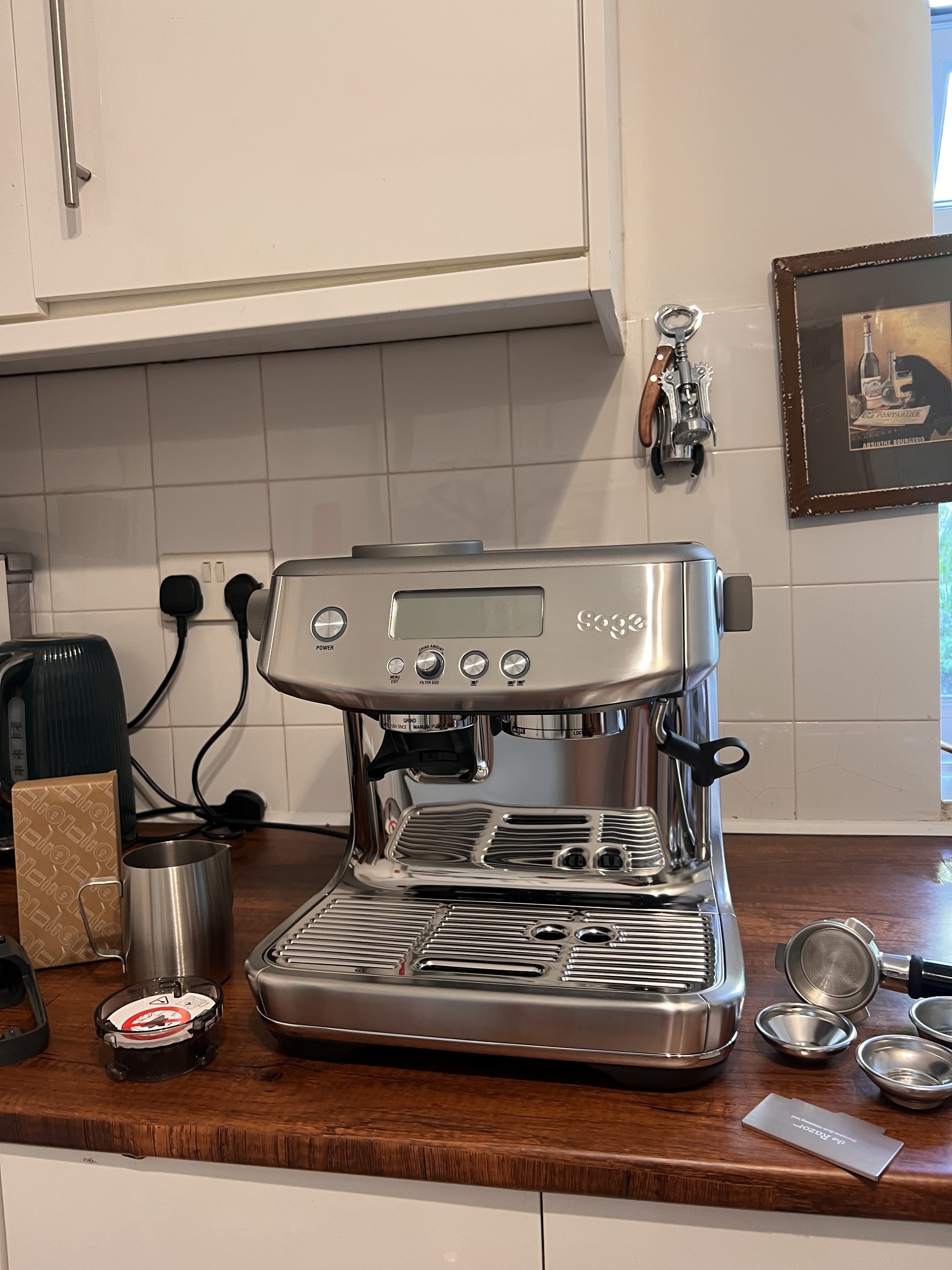 Image of a silver Sage Barista Pro machine on a wooden countertop in a white kitchen with different coffee machine accessories around it.