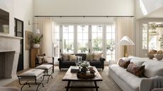 Transitional open-plan living room with large glass doors and sheer curtains, two sofas, a wooden coffee table, two wrought iron stools, a large stone fireplace, and a Persian neutral rug.