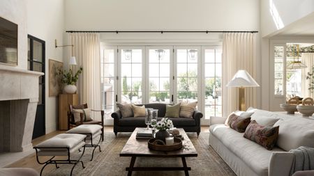Transitional open-plan living room with large glass doors and sheer curtains, two sofas, a wooden coffee table, two wrought iron stools, a large stone fireplace, and a Persian neutral rug.