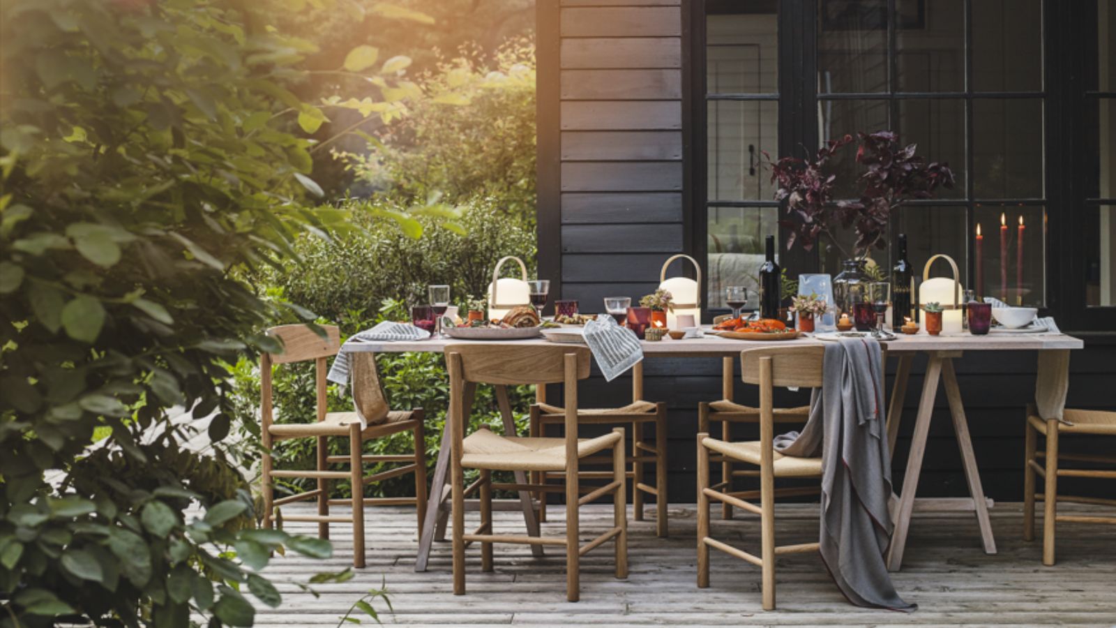 A patio with table and chairs laid for a meal outdoors, candles lit, place settings and glasses and dishes of cooked food