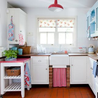 White kitchen with Belfast sink, terracotta floor tiles, floral blind and red light