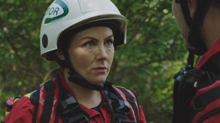 Stevie Nash in red HEMS uniform and crash helmet talking to Iain in a forest.