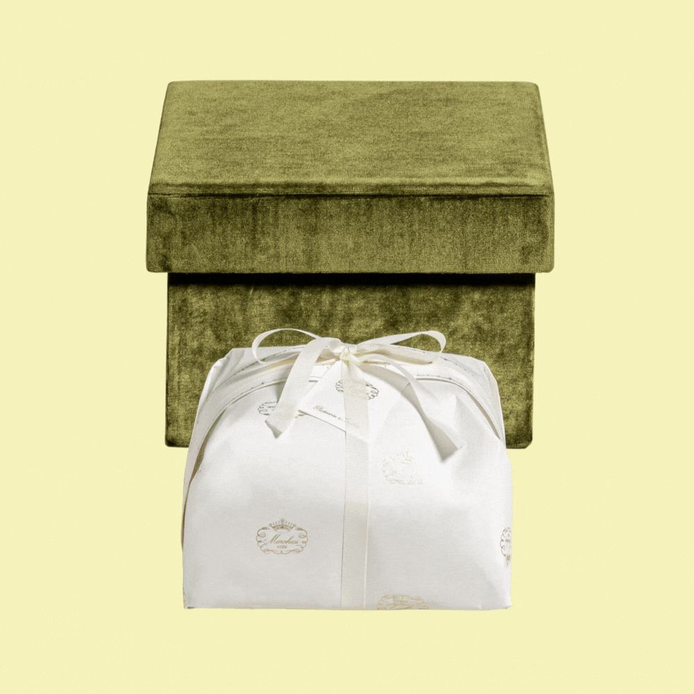 A green velvet box is captured behind a beautifully packaged white panettone with an elegant satin ribbon and, in a recurring gif, shown inside the box, decorated with floral motifs and placed against a pale yellow background.