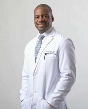 Dr Gbolahan Okubadejo is a man of color, pictured in a white doctor's coat, with a shirt and tie. He is pictured against a white professional backdrop for a headshot