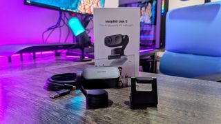 The Insta360 Link 2 webcam's packaging and included contents spread on a desk with violet backlighting, including the Insta360 Link 2, magnetic stand, and USB cable.