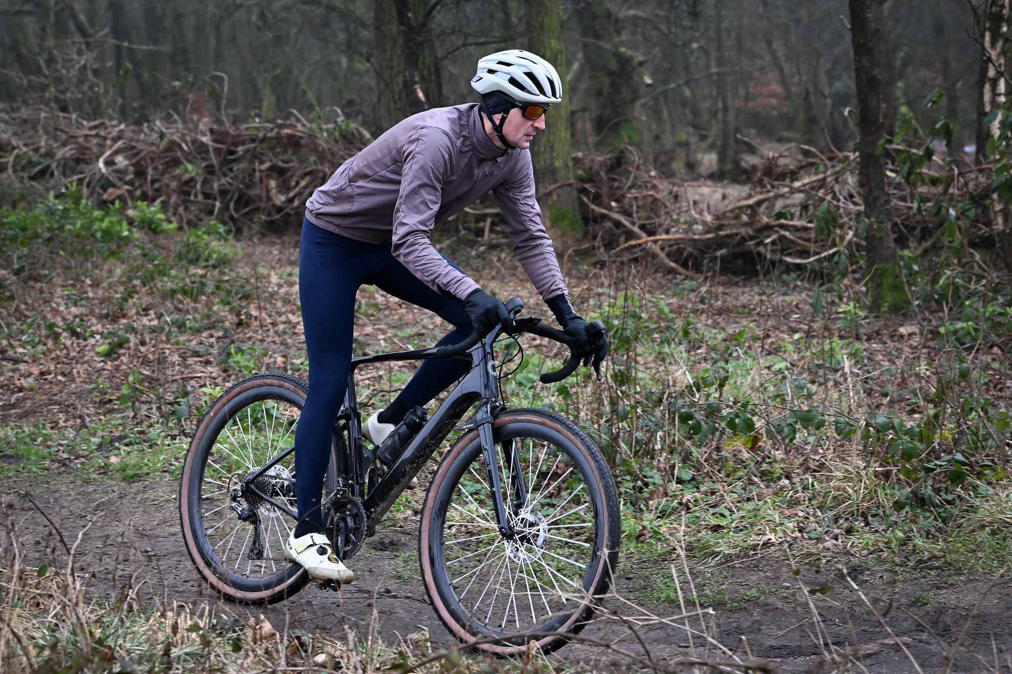 side view of a man wearing navy tights and purple jacket riding a black gravel bike on a dirt trail