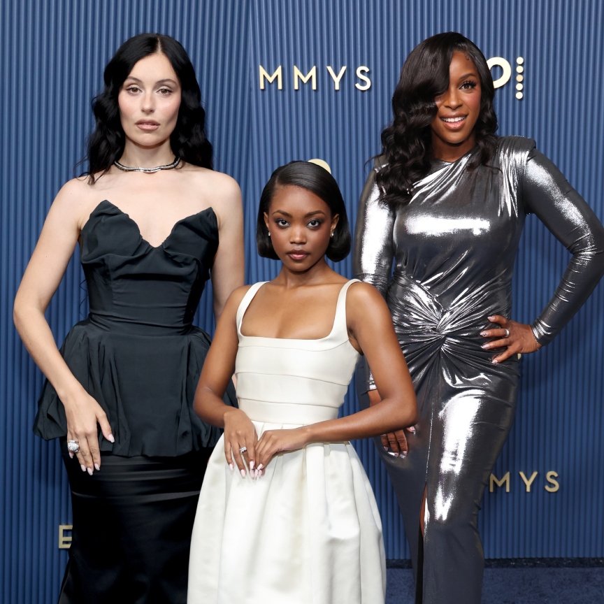 three women in glamorous hair, makeup, and gowns on a blue Emmys step and repeat