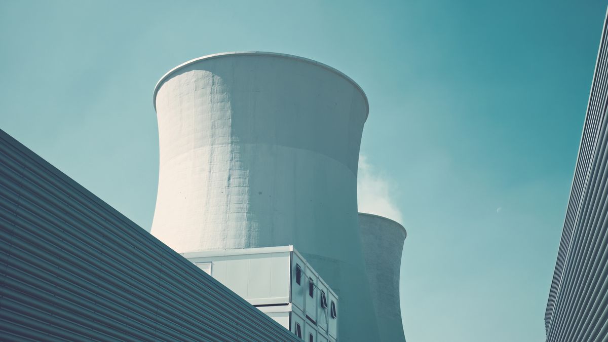 The cooling towers of a nuclear power plant photographed from the ground against a pale blue sky. The cooling towers are painted white and are framed to either side by corrugated steel structures.