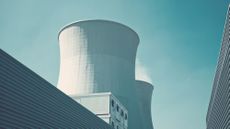 The cooling towers of a nuclear power plant photographed from the ground against a pale blue sky. The cooling towers are painted white and are framed to either side by corrugated steel structures.