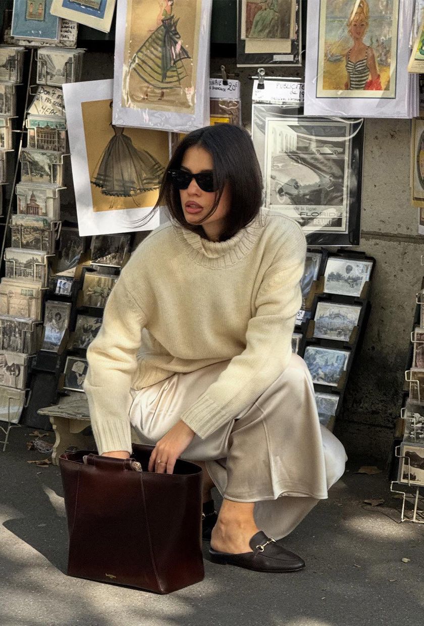 A woman wearing a white turtleneck with white satin pants, brown loafers, and a brown handbag.