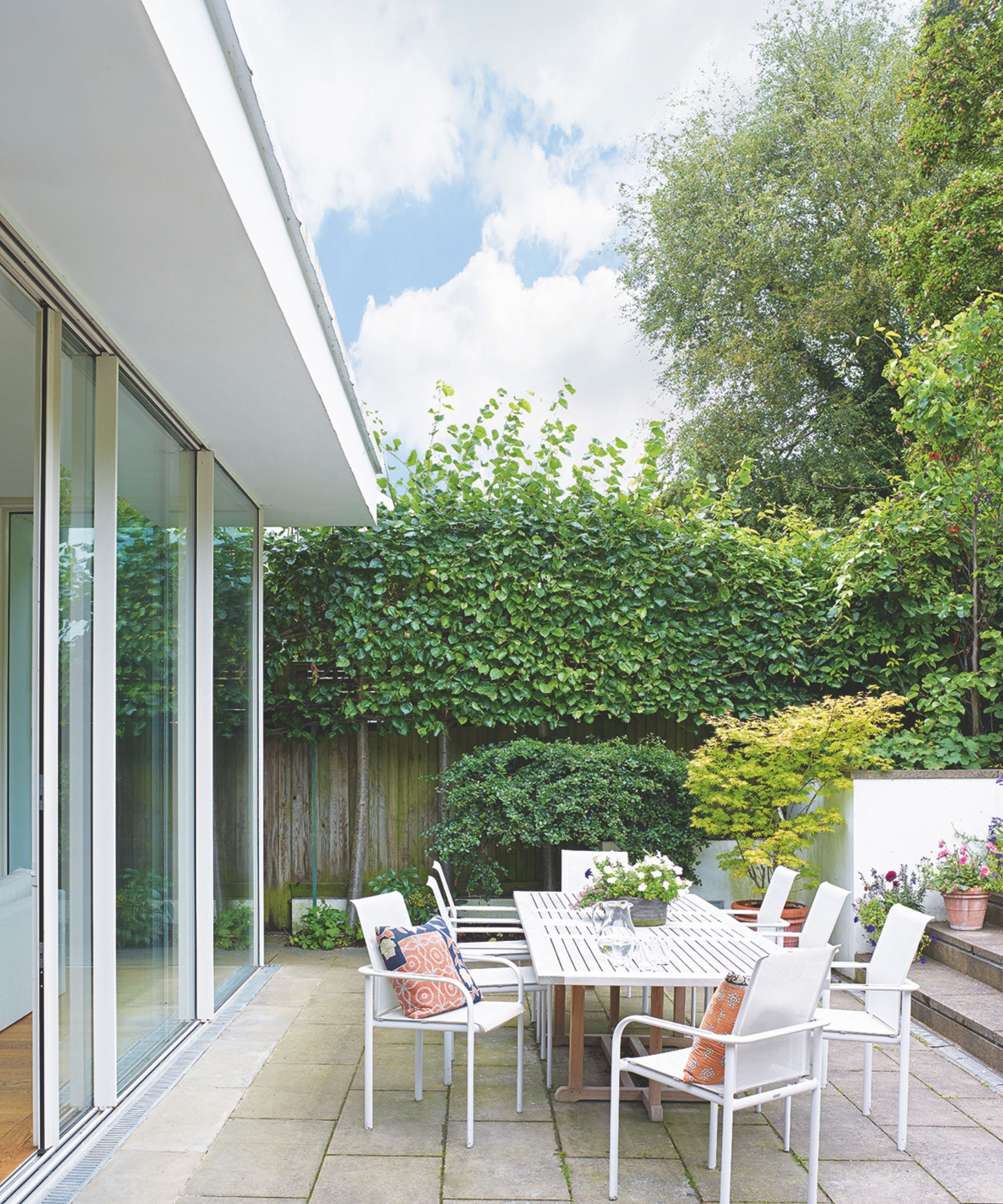 Modern house with glass doors leading out onto a patio with a white metal dining set including a long table and chairs, and privacy planting with hedges and trees.