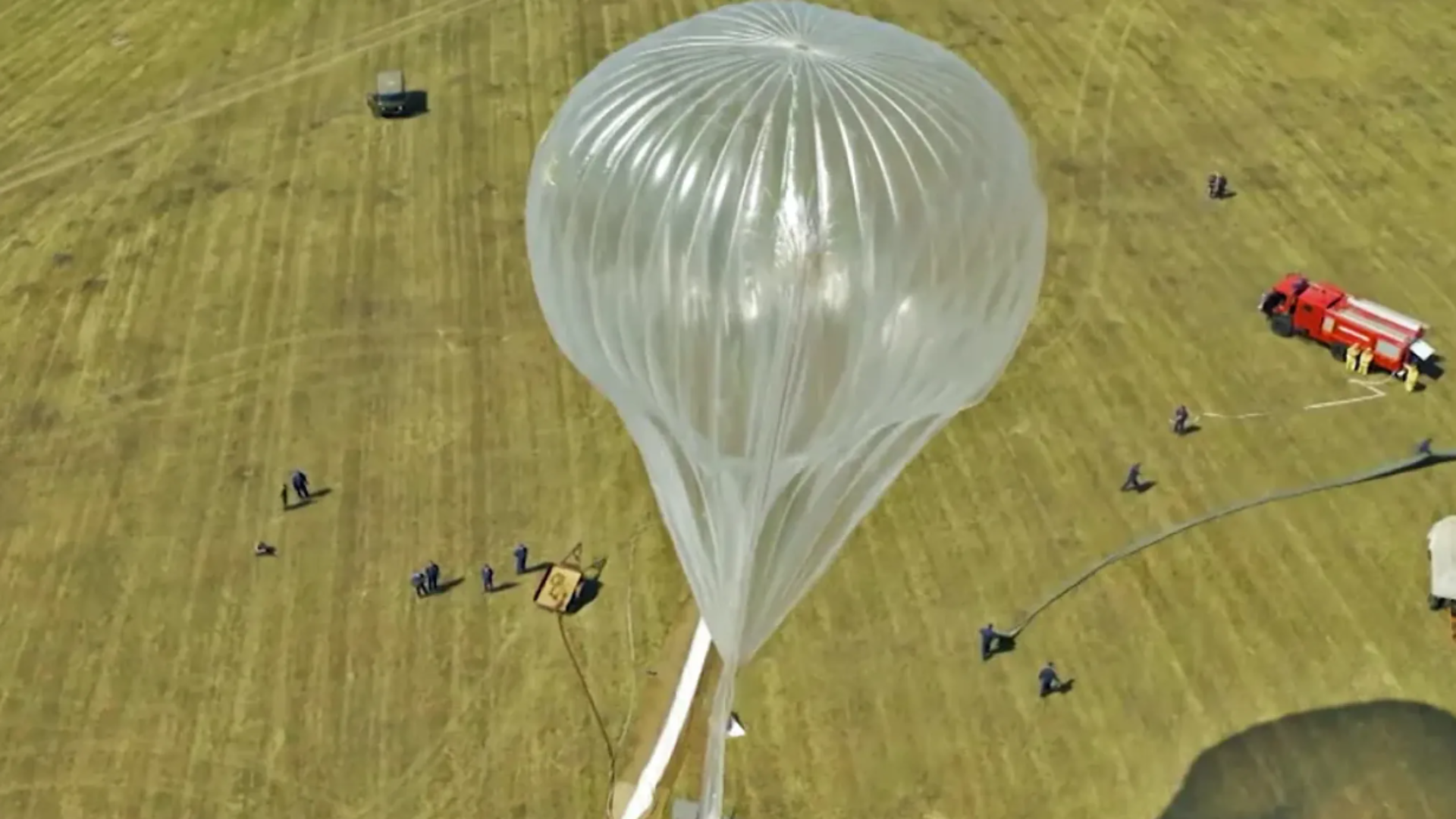 A Russian high-altitude stratospheric balloon is being prepared for launch in an open field, with ground crew and support vehicles positioned nearby.