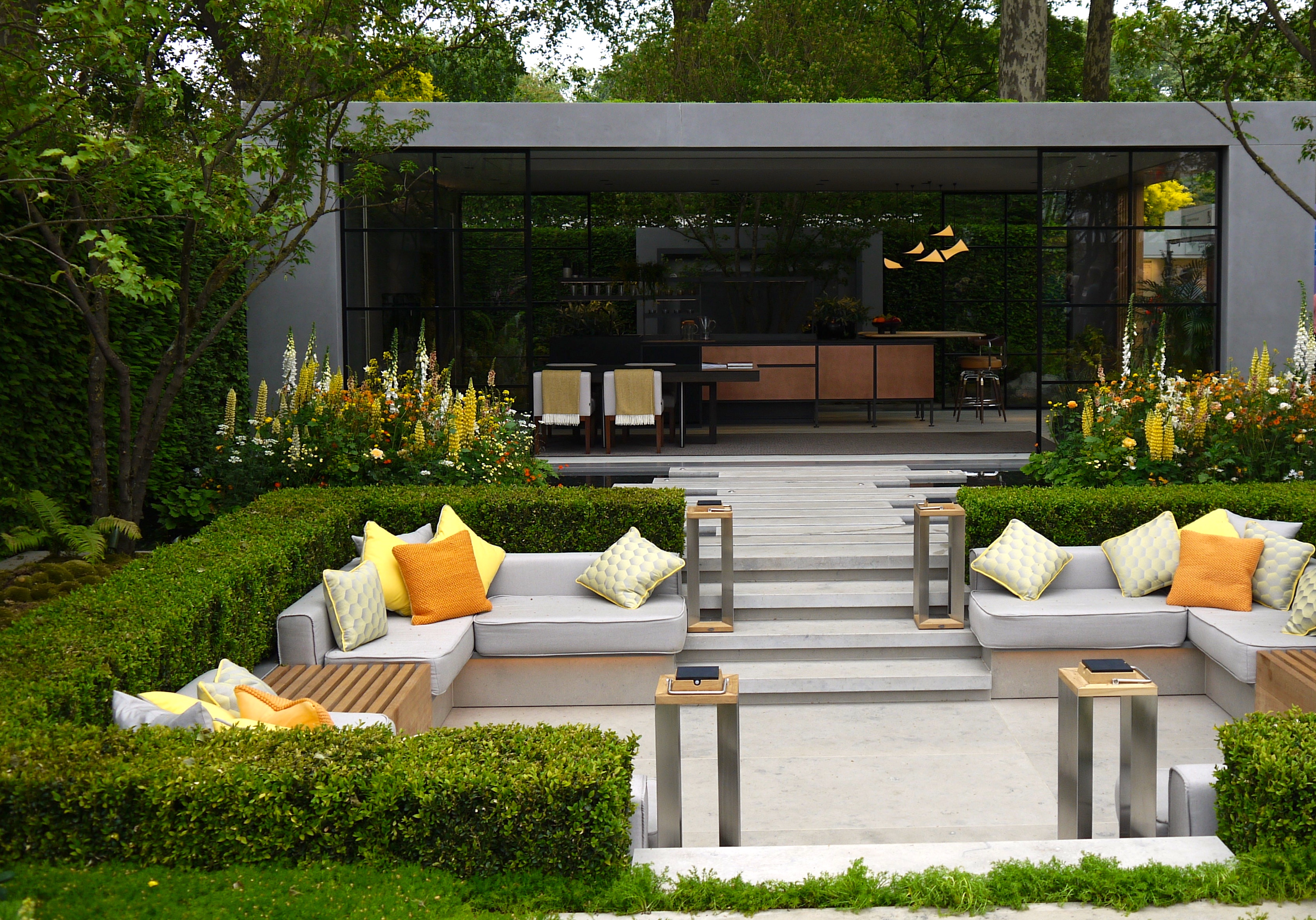 A sunken outdoor seating area surrounded by a neatly trimmed vibrant green hedge with yellow cushions and yellow flowers planted along its corners in front of the home's sliding doors