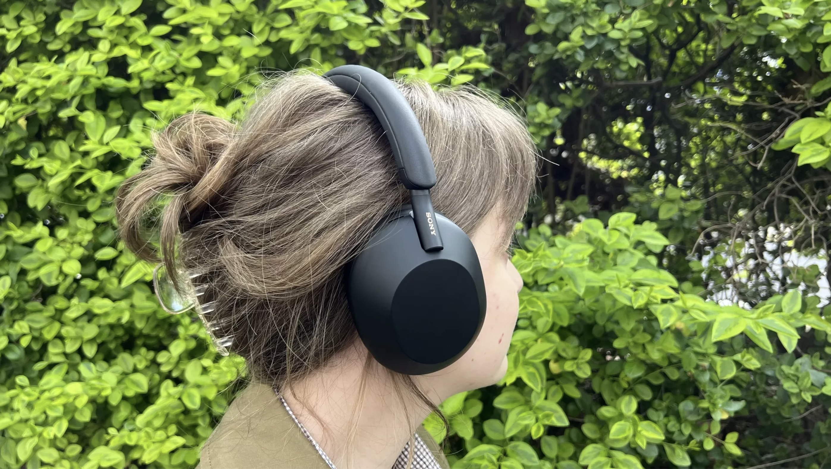 The Sony WH-1000XM5 wireless headphones in black being worn by a member of the TechRadar team outside in front of a green hedge.
