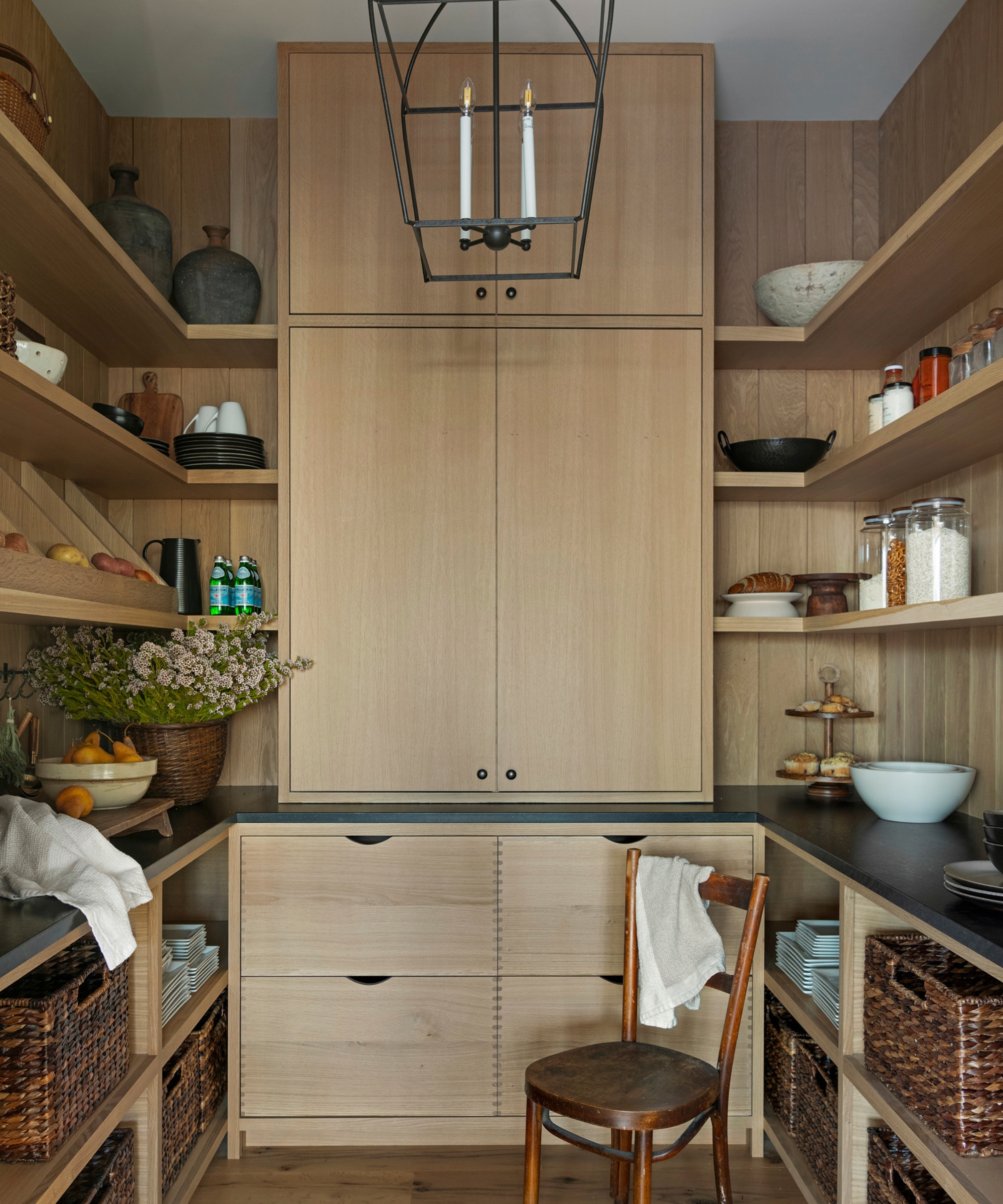 A pantry with white oak cabinetry and wall paneling, black countertops, and a mix of cabinet, drawer, and open storage