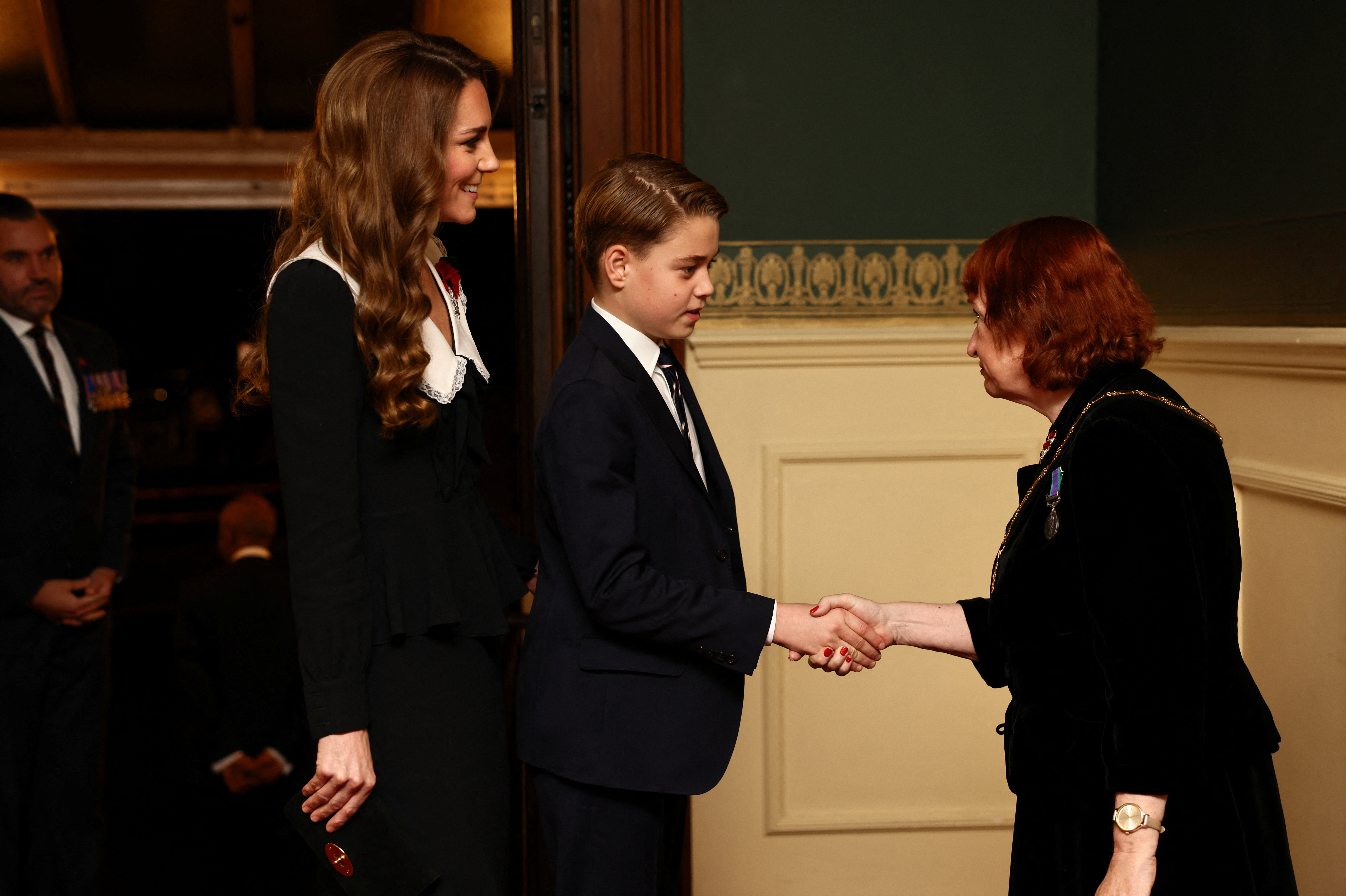 Prince George wearing a suit shaking hands with a woman with Princess Kate standing behind him