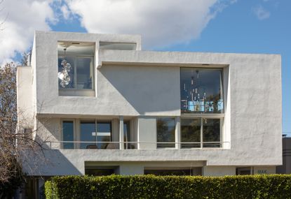 Rudolph M. Schindler apartment in Los Angeles, white rectangle shape building, windows, viewing balcony with silver hand rail, green hedge, tree branches to the left, blue sky with white clouds