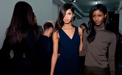 Room full of people and three female models wearing looks from the Ports 1961 collection. One model is wearing a black belted piece. Another model is wearing a dark blue V-neck dress with buttons. And the third model is wearing a brown turtle neck, chain and brown skirt