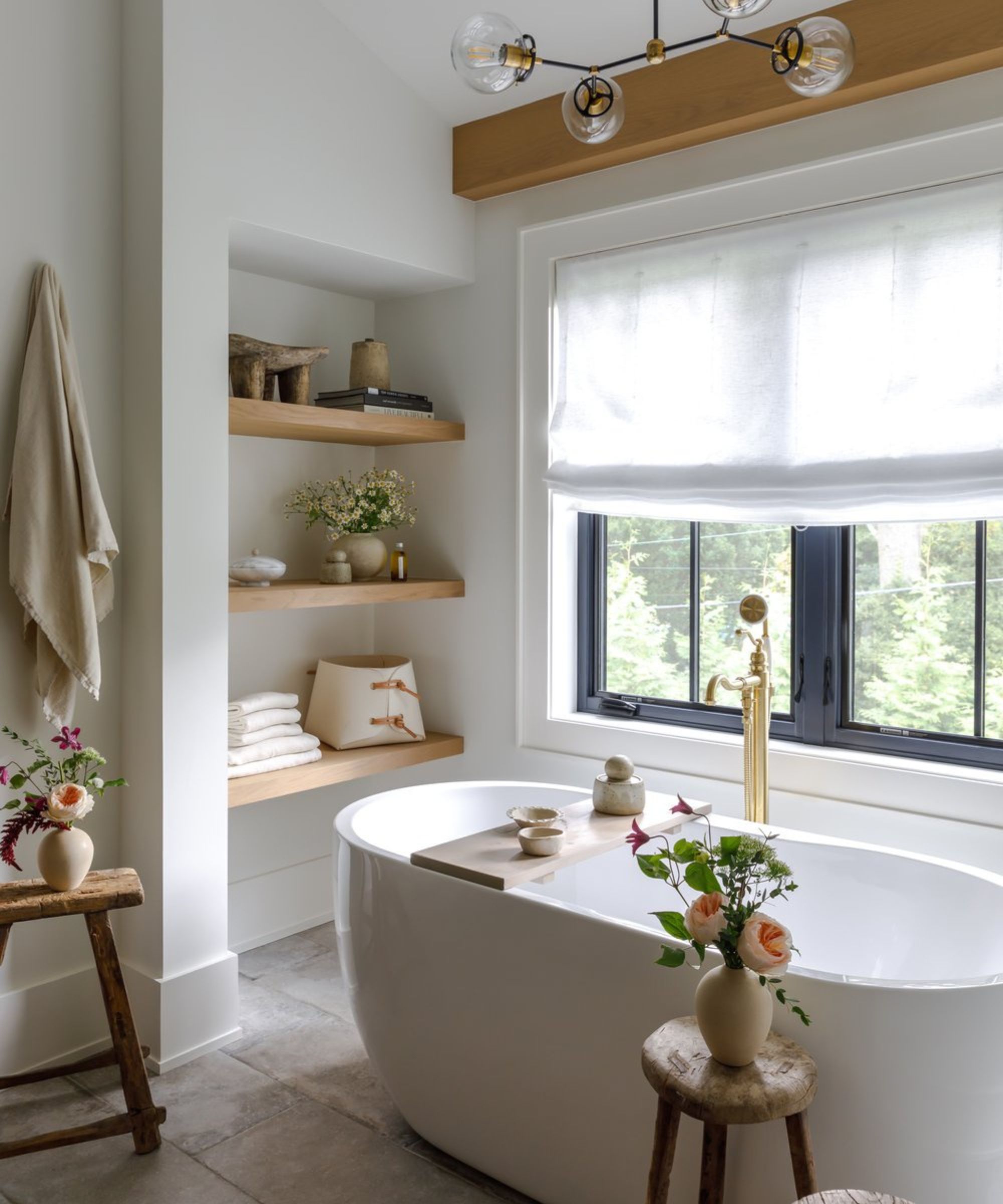 Small white spotless clean bathroom with freestanding bath and shelving. There are wooden stools with vases of flowers around, as well as bulbous statement ceiling light and linen window treatment