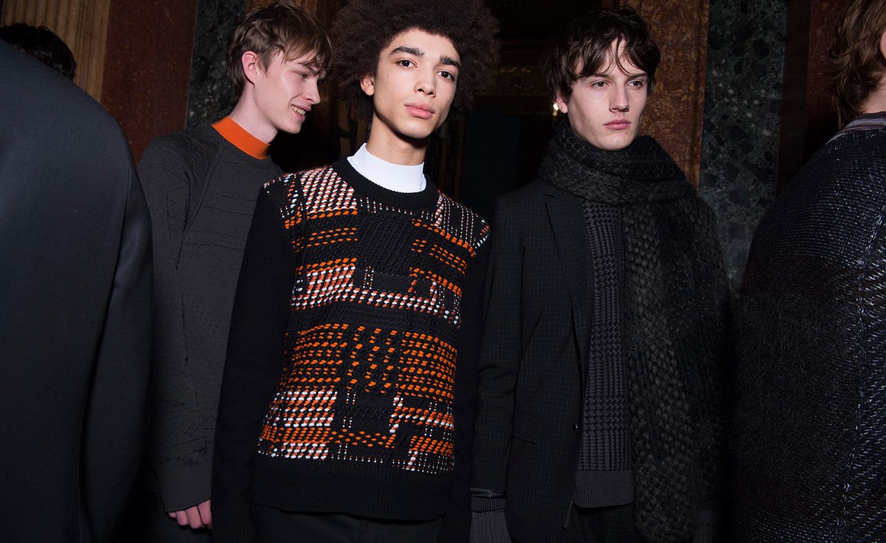 Three male models wearing looks from Pringle of Scotland&#039;s collection. One model is wearing a dark grey jumper with orange peaking above the neckline. Next to him is another model wearing a black, white and orange patterned jumper. And the third model is wearing a black jacket and black and grey patterned scarf