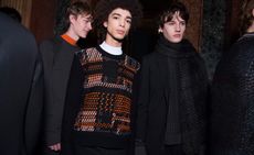 Three male models wearing looks from Pringle of Scotland's collection. One model is wearing a dark grey jumper with orange peaking above the neckline. Next to him is another model wearing a black, white and orange patterned jumper. And the third model is wearing a black jacket and black and grey patterned scarf