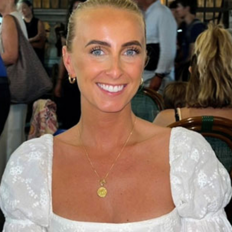 female with blonde hair pulled back off face wearing white top with gold necklace, people in background