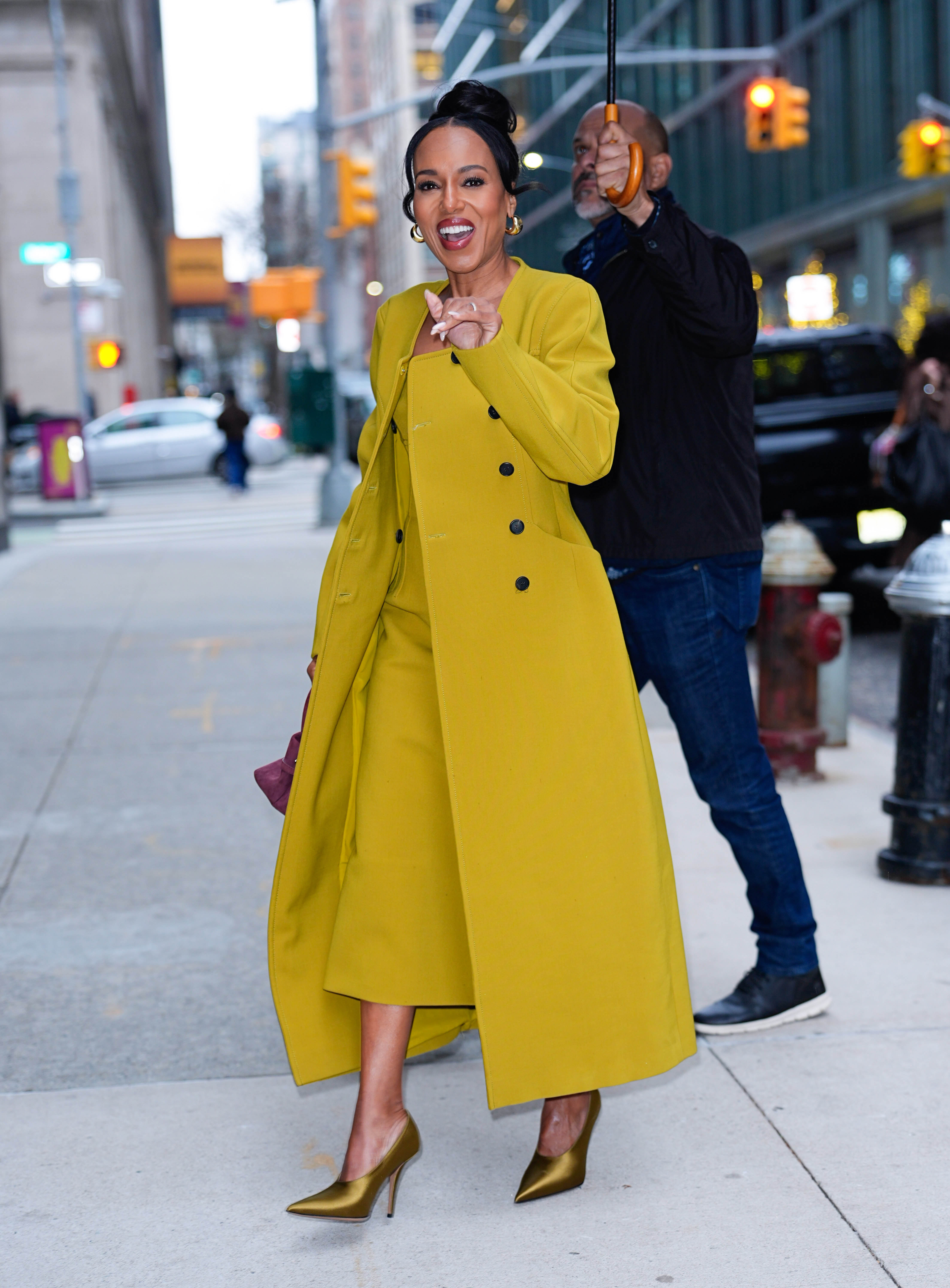 a paparazzi photo of a black woman wearing a long, mustard yellow coat and gold pumps