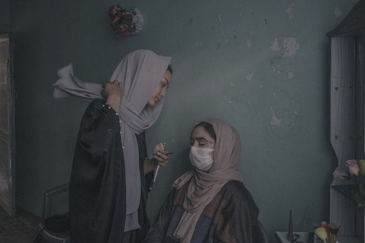 Two women in hijabs, one applying make-up on the other, inside a dimly lit room with a green wall and minimal decor