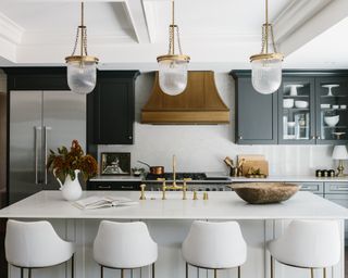 A kitchen with white marble topped island, white upholstered bar stools, black kitchen cabinets, bronze hood and pendant lights